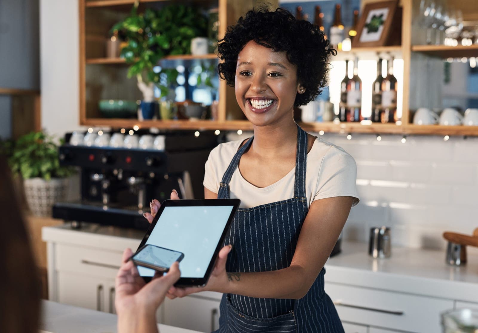 A smiling barista holding a tablet as a customer pays with a phone