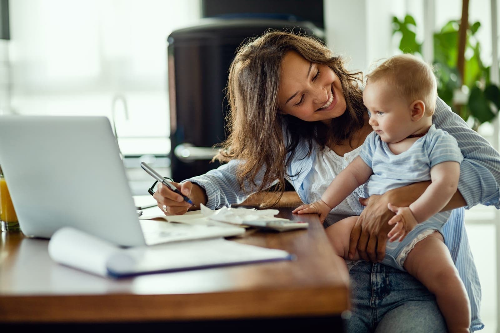 A mother holding her baby while working through bills at a laptop